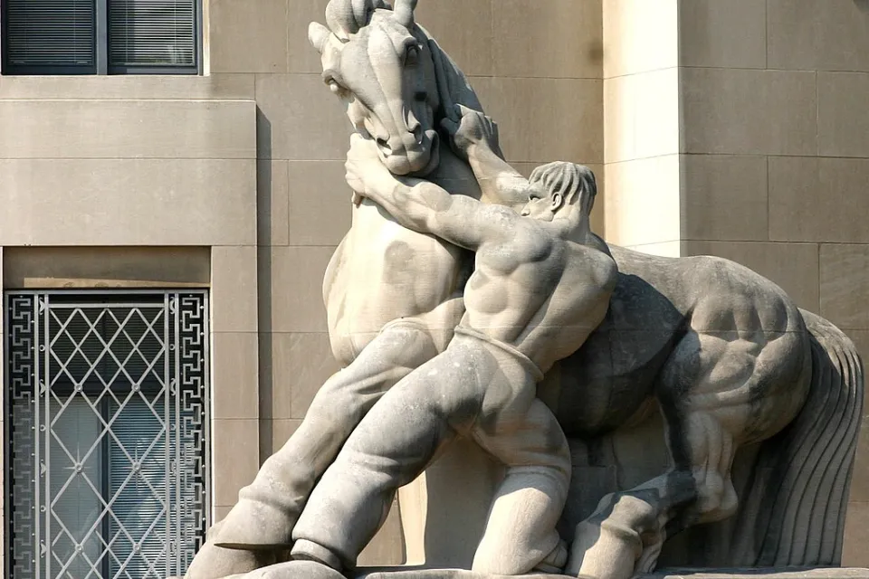 A stone statue of a shirtless muscular man wrestling with a powerfully built horse, his hands on the horse's neck, pulling downward.